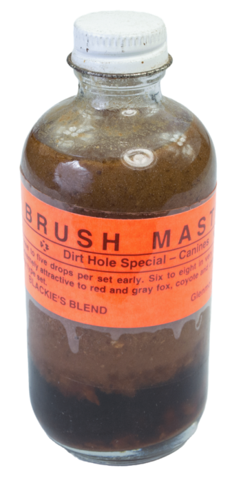 A small glass bottle labeled "Blackie's Brush Master Lure" is filled with brown liquid and sealed with a metal cap.